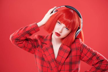 Kırmızı dudaklı, kızıl saçlı, kırmızı kareli ceket ve kulaklık takan şık bir kız. DJ, parti kızı. Kırmızı stüdyo geçmişi. Güzellik, Moda. 