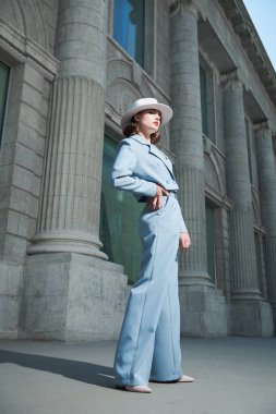 Hollywood tarzı. Güzel kahverengi saçlı, zarif bir gökyüzü mavi takım elbiseli ve beyaz şapkalı bir kadının şehir caddesinde durduğu uzun bir kare. 40 'lı yılların tarzı. Makyaj ve saç stili.