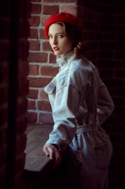 A beautiful young woman with elegant makeup and hairstyle dressed in denim overalls and red beret stands by the window in a room with brick walls. Beauty, fashion. Retro style.