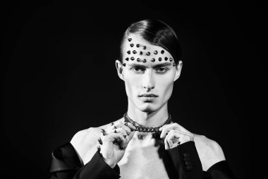 Stüdyoda, siyah ceketli yakışıklı bir erkek mankenin yüzünde ve elinde metal çivilerle poz verirken çekilmiş. Karanlık arka plan. Siyah beyaz portre.