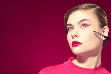 Parlak pembe rujlu ve gözlerinde pembe oklar olan güzel bir model kız fuşya arka planında fırçayla makyaj yapıyor. Güzellik, makyaj ve kozmetik. Makyaj aletleri. Boşluğu kopyala.