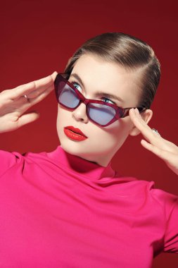 Parlak makyajlı ve gözlüklü zarif sarışın kız kırmızı arka planda poz veriyor. Makyaj ve makyaj malzemeleri. Gözlük stili. 