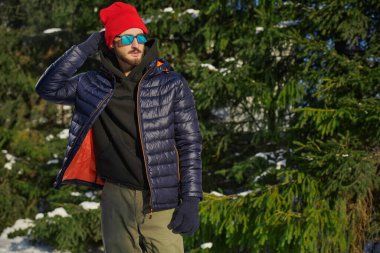Kışlık ceketli yakışıklı bir esmer adam portresi örgü bir şapka ve güneş gözlüğü kış parkında karla kaplı köknar ağaçlarının yanında duruyordu. Kış modası. Boşluğu kopyala.