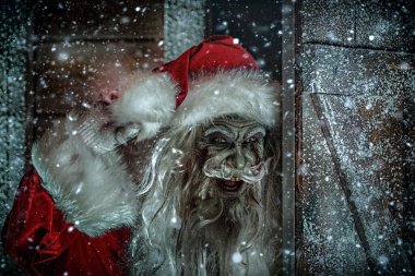 Korkunç bir Noel Baba zombisinin portresi. Tahta bir kapının arkasından sokağa bakıyor ve gülüyor. Kar yağışı. Noel 'de kabus. Cadılar Bayramı. 