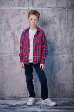 Ekose gömlek ve kotla arka planda duran ve gülümseyen şık saç stili olan yakışıklı bir çocuğun tam boy portresi. Çocuk modası.