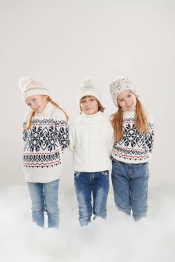 Kış çocukları. Kotlu, kalın örülü kazaklı ve şapkalı üç neşeli arkadaş kar yığınında yan yana durur ve gülümserler. Çocuk modası. Kış aktiviteleri.
