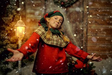 Mutlu noeller. Neşeli küçük bir elf kızı kar altında, kış ormanındaki ahşap bir kulübede duruyor. Noel Masalı.