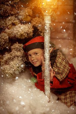 Neşeli küçük bir elf kızı, karlı bir gecede yanan bir fenerin altında ahşap bir evin yanında oturur ve gülümser. Noel Masalı. 