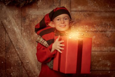 Mutlu noeller. Neşeli küçük bir elf kızı elinde kırmızı bir hediye kutusuyla kış ormanındaki ahşap bir kulübede duruyor. Noel hazırlıkları ve hediyeleri.