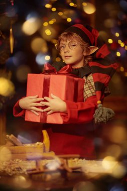 Küçük sevimli elf çocuk Noel için marangoz atölyesinde çocuklar için ahşap oyuncaklar yapıyor ve onları hediye kutularına koyuyor. Noel Baba atölyesi. Sihirli ışıklar yanıp sönüyor. Noel Masalı.