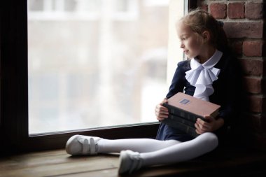 Okul üniformalı küçük bir kız pencerenin kenarında oturuyor ve kucağında kitaplarla pencereden dışarı bakıyor. Doğal ışıklandırma. Eğitim. Çocuklar, çocukluk, duygular. 
