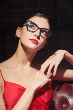 Zeki ve zarif. Akşam makyajlı, kırmızı saten elbiseli ve deri koltukta oturan zarif gözlüklü güzel bir kızın portresi. Giysiler ve aksesuarlar içinde şık bir akşam stili..