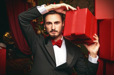 Pahalı bir gece elbisesi içindeki şık esmer adam elinde kırmızı bir hediye kutusu tutuyor. Tatil modası. Alışveriş ve hediyeler. Noel içi hediye kutularıyla dolu.. 
