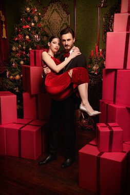 Zarif takım elbiseli yakışıklı bir adam kırmızı gece elbisesi içindeki sevgilisini kollarında tutuyor. İçinde bir sürü hediye kutusu olan lüks bir Noel içeceği. Noel ve Yeni Yıl kutlaması. Tatil modası. 