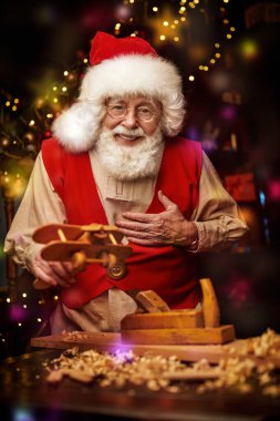 Mutlu gülümseyen Noel Baba atölyesinde çocuklar için parlak ışıklarla süslenmiş tahta oyuncaklar yapıyor. Noel gelenekleri ve peri masalları.