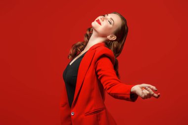 Şık, kırmızı ceketli, kollarını zevkle yana açan zarif makyajlı, genç bir kadının stüdyo portresi. Kırmızı arka plan. Mutluluk ve başarı.