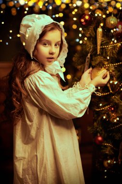 Magic Christmas. A pretty little girl in a nightie and a nightcap admires the beauty of the decorations hanging on the Christmas tree. Vintage style.