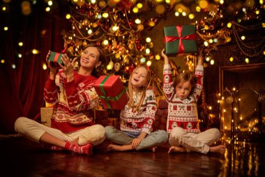 Güzel bir anne ve tatlı kızları, Noel ağacı ve şöminenin yanında oturmuş, hediyelerle seviniyorlar. Hoş, güzel dekore edilmiş bir oda. Mutlu Noeller. Noel kutlaması ve hediyeler.