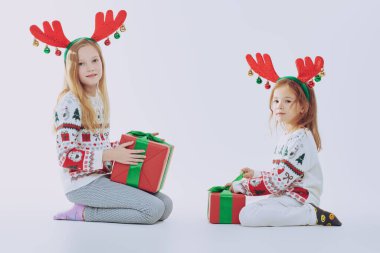 Noel süveteri ve geyik boynuzu giymiş iki tatlı kız Noel hediyeleriyle karşılıklı oturup gülümsüyor ve gülüyor. Konfetili beyaz arka plan. Kış çocuğu modası. Noel ve Yeni Yıl.