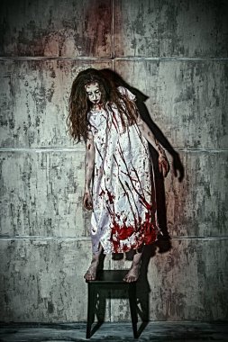 Kana bulanmış bir kadının, karanlık bir odada, bir taburede durduğu korkunç bir sahne. Kana susamış zombi kadın. Korku filmi, gerilim. Cadılar Bayramı.