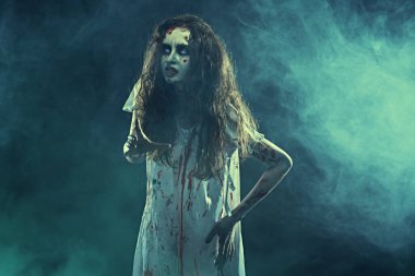 Ürkütücü zombi kadın. Korkunç şekilsiz, solgun gözlü bir kadın, şeytan tarafından ele geçirilmiş, kanlı bir gecelik kıvrılmış, karanlıkta duruyor, dumanla çevrili. Korku, gerilim. Cadılar Bayramı.