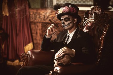 Dia de los muertos. Ölüler Günü. Zarif ve tehlikeli Baron Samedi eski bir kalede puro içiyor. Kafatası şekeri makyajlı yakışıklı adam. Cadılar Bayramı.