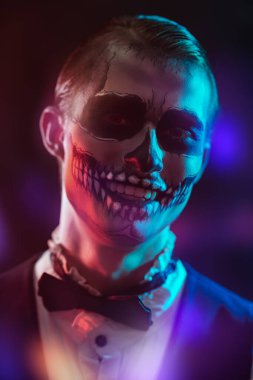 Kafatası makyajlı yakışıklı zarif bir adam siyah arka planda neşeli renkli bir ışıkla gülümsüyor. Dia de los muertos. Ölüler Günü. Cadılar Bayramı.