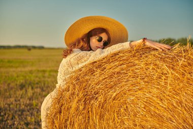 Modern boho, hippi tarzı. Örgü kazaklı, geniş kenarlı hasır şapkalı ve batan güneşin altında samanlıkta duran yuvarlak gözlüklü manken. Sonbahar.