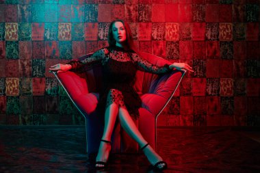 Siyah bir gece elbisesi içinde lüks dairelerde koltukta oturan güzel bir kadının tam boy portresi. Göz kamaştırıcı bir yaşam tarzı. Güzellik, moda. Modern iç mekan, mobilya.