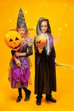 Cadılar Bayramı büyüsü. Cadılar Bayramı balonu ve balkabağı sepeti tutan bir cadı ve büyücü kostümü giymiş iki mutlu çocuk. Sarı arka planda sihirli ışıklar var. Şaka mı şeker mi?.