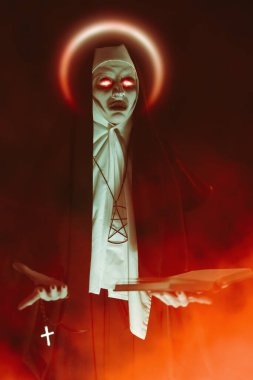 Halloween. Horrors. An evil cursed nun with red burning eyes holds a bible and a cross in her hands standing on a black background with red haze.  clipart