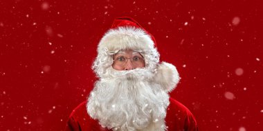 Noel Baba kameraya şaşkınlıkla bakıyor. Kırmızı stüdyo arka planı ve kar yağışı. Uzayı kopyala Noel ve Yeni Yıl.