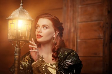 Peri masalı kahramanı. Güzel kızıl saçlı bir peri prensesi, ahşap bir evin arka planında fenerin ışığına büyülenmiş gibi görünür. Boşluğu kopyala.