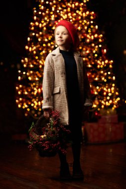 Çocuk modası ve Noel. Kırmızı bereli ve ekose ceketli güzel bir kızın tam boy portresi, elinde bir sepetle şenlik ortamında duruyor ve mutlu bir şekilde gülümsüyor..