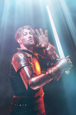 Yükseltilmiş kılıçlı yakışıklı bir ortaçağ şövalyesinin portresi, yukarıdan gelen ilahi ışıkla aydınlanan, karanlık bir arka planda duruyor. Bir şövalye romanının kahramanı.. 