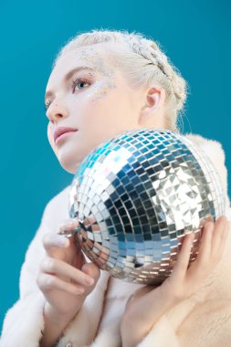 Kış tatili güzeli. Hafif makyajlı, yüzünde parıltılarla beyaz kürklü, beyaz kürklü ve aynalı disko topuyla poz veren güzel bir kız. Mavi stüdyo arka planı. 