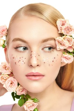 Kozmetik ve makyaj malzemeleri. Açık renk makyajı, parlak çilleri ve soluk pembe gülleri olan narin sarışın bir kızın yakın plan portresi. Güzellik konsepti. Beyaz arka plan. Güzellik ürünleri. Parfüm.