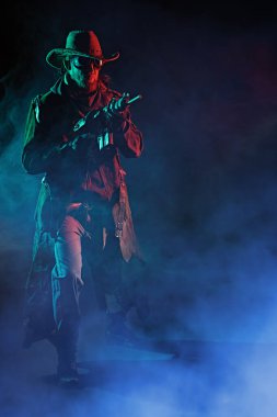 Vahşi Batı tarzında giyinmiş, gözlüklü ve tüfekli bir adam kıyametten kurtuldu. Stüdyo portresi. Sis dolu siyah bir arkaplan. Kıyameti haber veren kovboy.. 