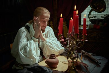 Geniş kollu beyaz gömlekli yakışıklı bir asilzade aile malikanesinde mum ışığında bir masada oturuyor ve mektubunu düşünüyor. 16.-17. yüzyılın tarihi yeniden yapılandırması. 