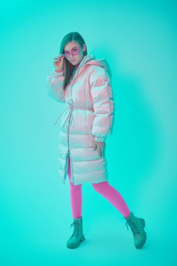 Kış kıyafetleri tarzı. Şık, uzun beyaz ceketli, parlak fuşya gözlüklü ve taytlı güzel bir kızın tam boy portresi. Aquamarine stüdyo geçmişi. 