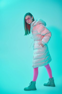 Kış kadın modası. Şık, uzun beyaz ceketli, parlak fuşya gözlüklü ve taytlı güzel bir kızın tam boy portresi. Mavi stüdyo arka planı. 