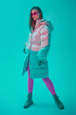 Yeni kış koleksiyonu elbiseler ve aksesuarlar. Uzun beyaz ceketli, parlak fuşya gözlüklü ve taytlı bir manken kızın tam boy portresi. Stüdyo aquamarine arkaplanı.