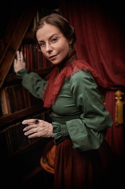 Eski moda iç mekan. Ev kıyafetleri ve gözlüklü zarif bir aristokrat bayan bir kitaplığın yanında duruyor. 19. yüzyıl - 20. yüzyıl başlarındaki romanın kahramanı..