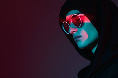 Gözlük stili. Siyah hırkalı, kapüşonlu ve zarif yuvarlak gözlüklü bir erkek mankenin portresi. Stüdyo, karışık renkli ışıklandırmayla çekildi. Boşluğu kopyala. 