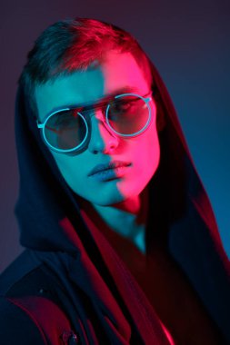 Siyah hırkalı, kapüşonlu ve zarif yuvarlak gözlüklü bir erkek mankenin portresi. Stüdyo, karışık renkli ışıklandırmayla çekildi. Gözlük stili.