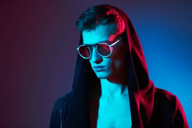 Siyah hırkalı, kapüşonlu ve zarif yuvarlak gözlüklü bir erkek mankenin portresi. Stüdyo, karışık renkli ışıklandırmayla çekildi. Gözlük stili. Boşluğu kopyala. 