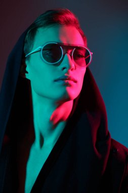 Moda fotoğrafı. Gözlük stili. Kapüşonlu ve şık gözlüklü yakışıklı genç bir adamın portresi stüdyoda karışık renkli ışıklandırmayla poz veriyor..