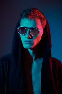 Siyah hırkalı, kapüşonlu ve zarif yuvarlak gözlüklü bir erkek manken. Stüdyo, karışık renkli ışıklandırmayla çekildi. Gözlük stili.