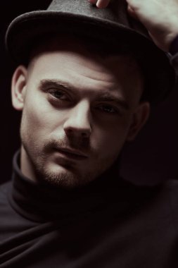 Siyah balıkçı yaka kazaklı ve kendine güvenen şapkalı yakışıklı bir adamın portresi. Stüdyo siyah arka plan. Siyah tuşlu portre.