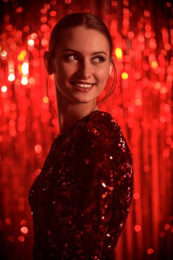 Gece makyajı ve parlak pullu gece elbisesiyle kırmızı folyo perdelere poz veren güzel bir kadının portresi. Şenlikli bir akşam stili. Noel ve Yeni Yıl.